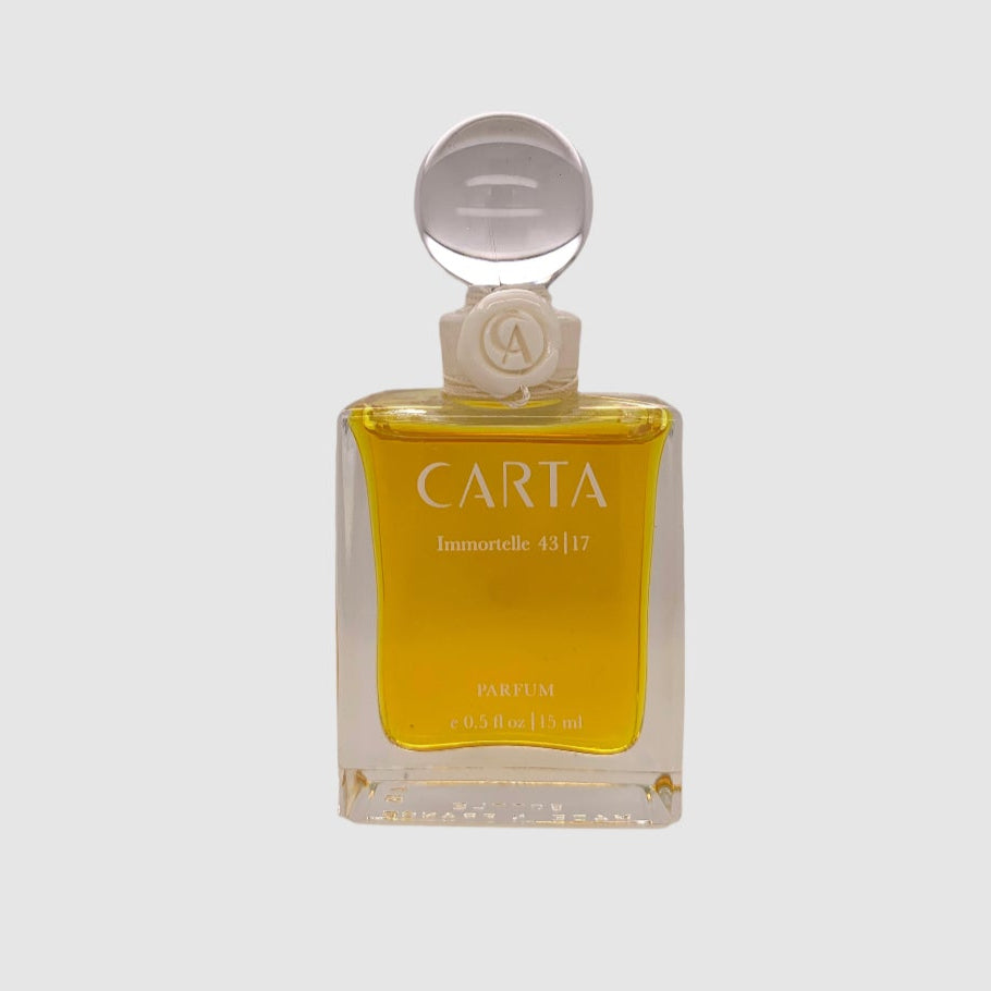 A clear glass perfume bottle with a yellow liquid inside, labeled 'CARTA parfums', with a white cap and a decorative element on the front.