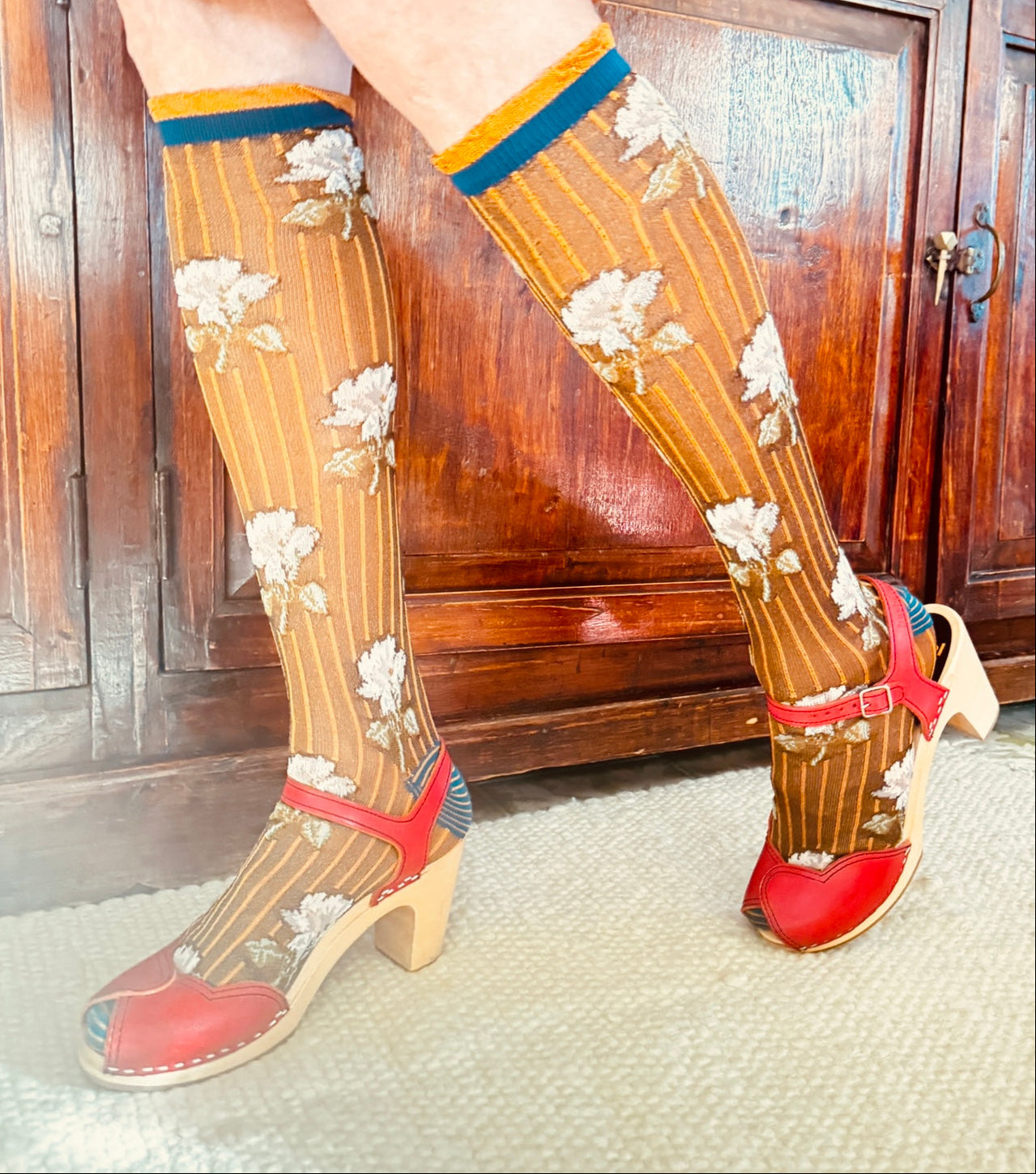 Legs wearing floral knee-high socks with red shoes in front of a wooden cabinet.