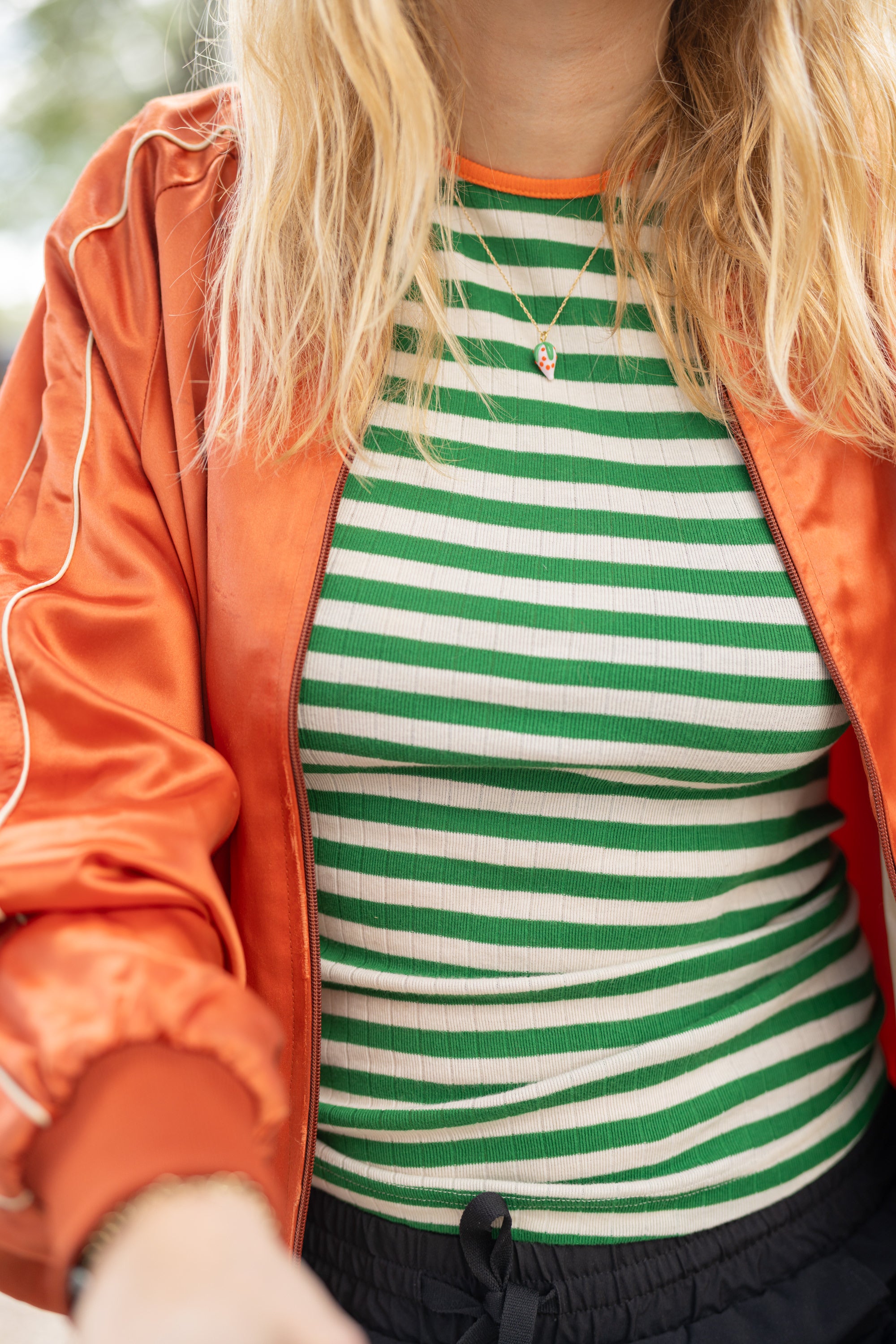 Person wearing a green and white striped shirt with an orange jacket and gold  necklace with glass strawberry charm