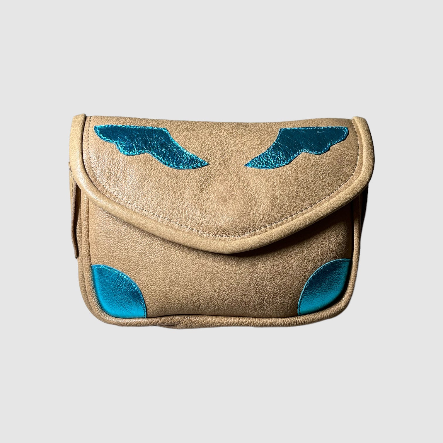 Small, soft, sand colored leather crossbody bag with metallic turquoise wing detail and accent applique. Front view.