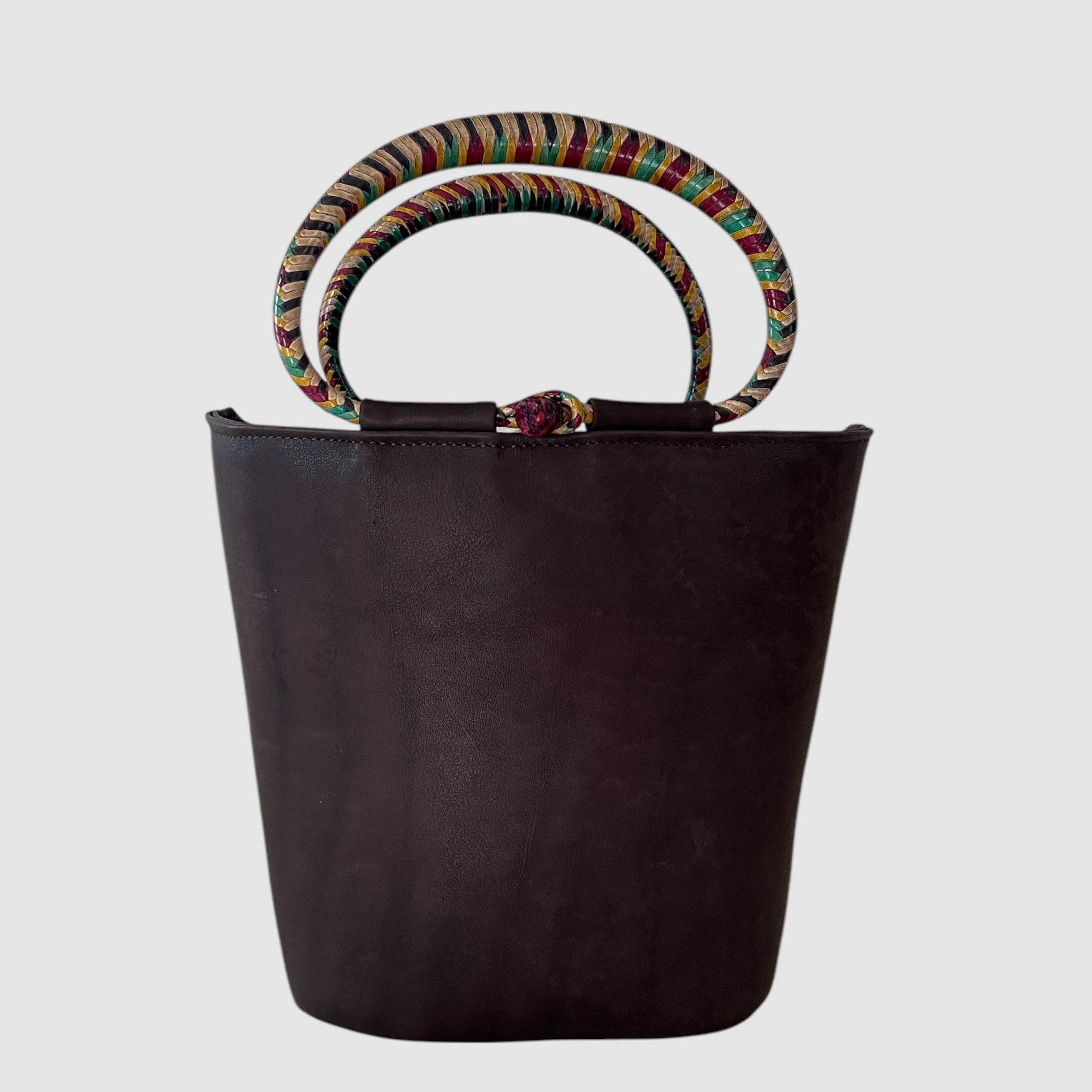 A matte chocolate brown leather bucket bag with two multi-colored hand woven leather handles and a zipper closure.