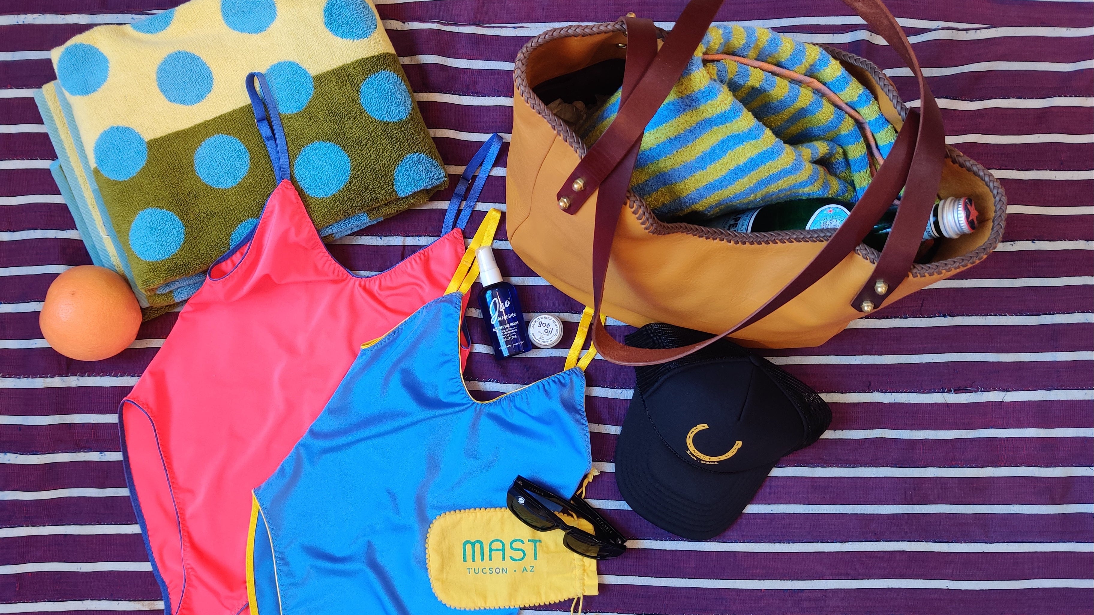 A styled flat-lay of beach and pool accessories on a purple and white striped blanket, featuring a tan tote bag, a blue and pink one-piece swimsuit, a yellow and blue polka dot towel, a blue and yellow striped towel, a black cap, sunglasses, and sunscreen.