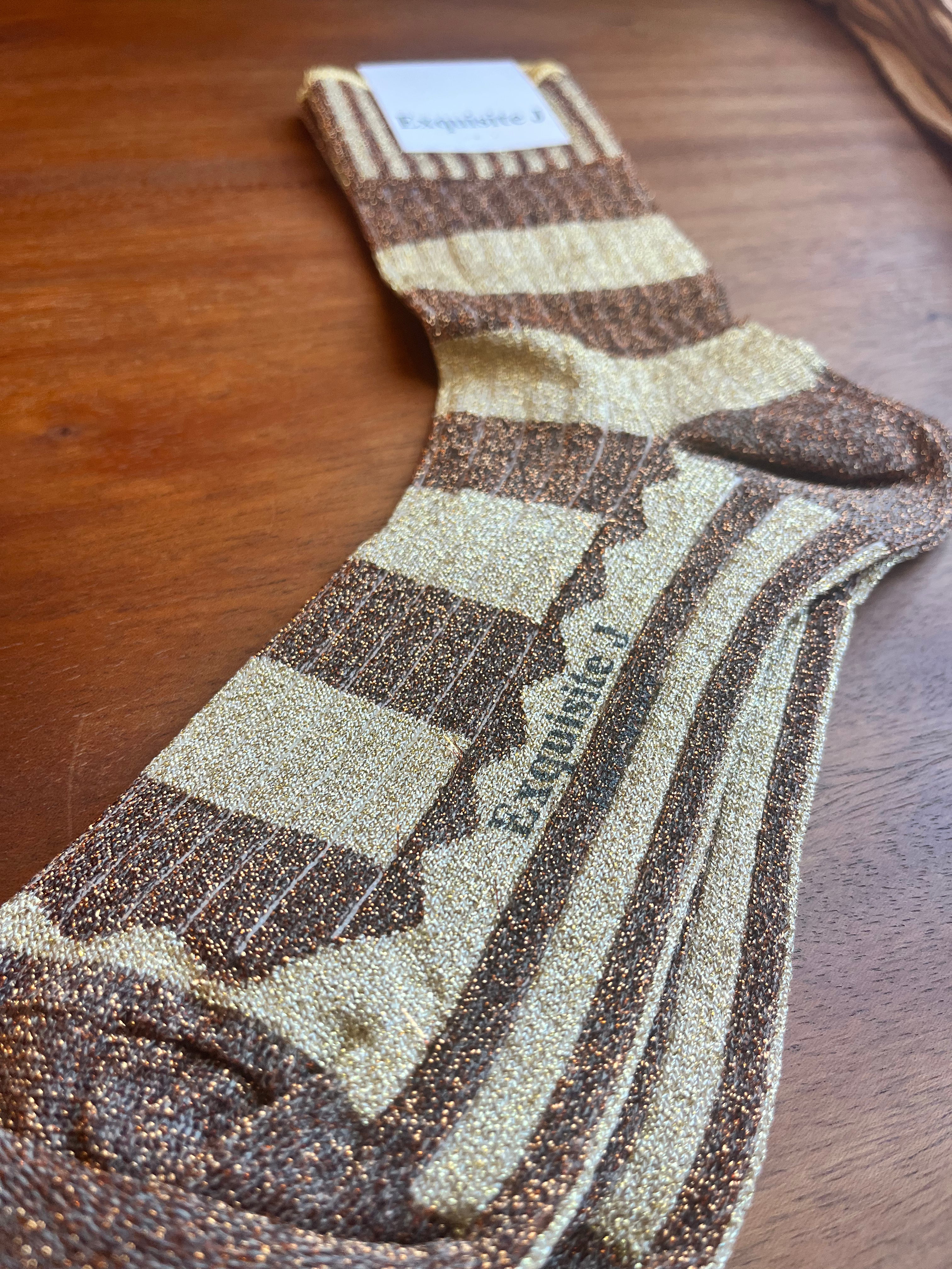 A pair of glittery striped socks in brown and beige, laid flat on a wooden surface. The brand name 'Exotique' is printed on the side of one sock, and a white brand tag is visible at the cuff.