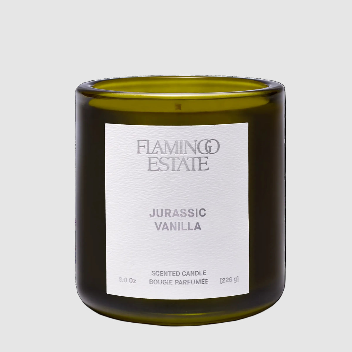 A scented candle in an olive green glass jar with a white paper label reading 'Flaming Estate Jurassic Vanilla' on a clean white background.
