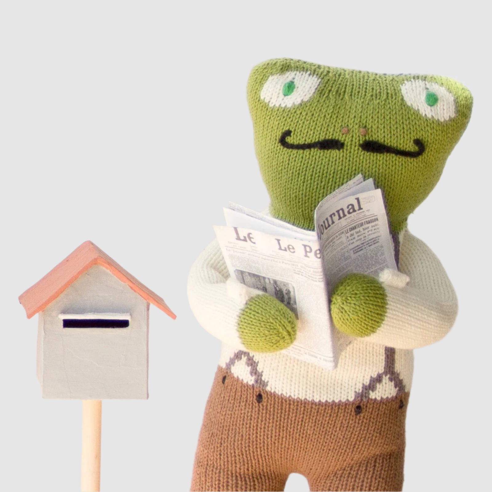 Knitted frog character reading a newspaper next to a mailbox on a light background