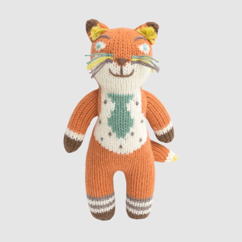 Knitted doll resembling a fox with a multi-colored design, featuring an orange body, green tie on the chest, and beige details.
