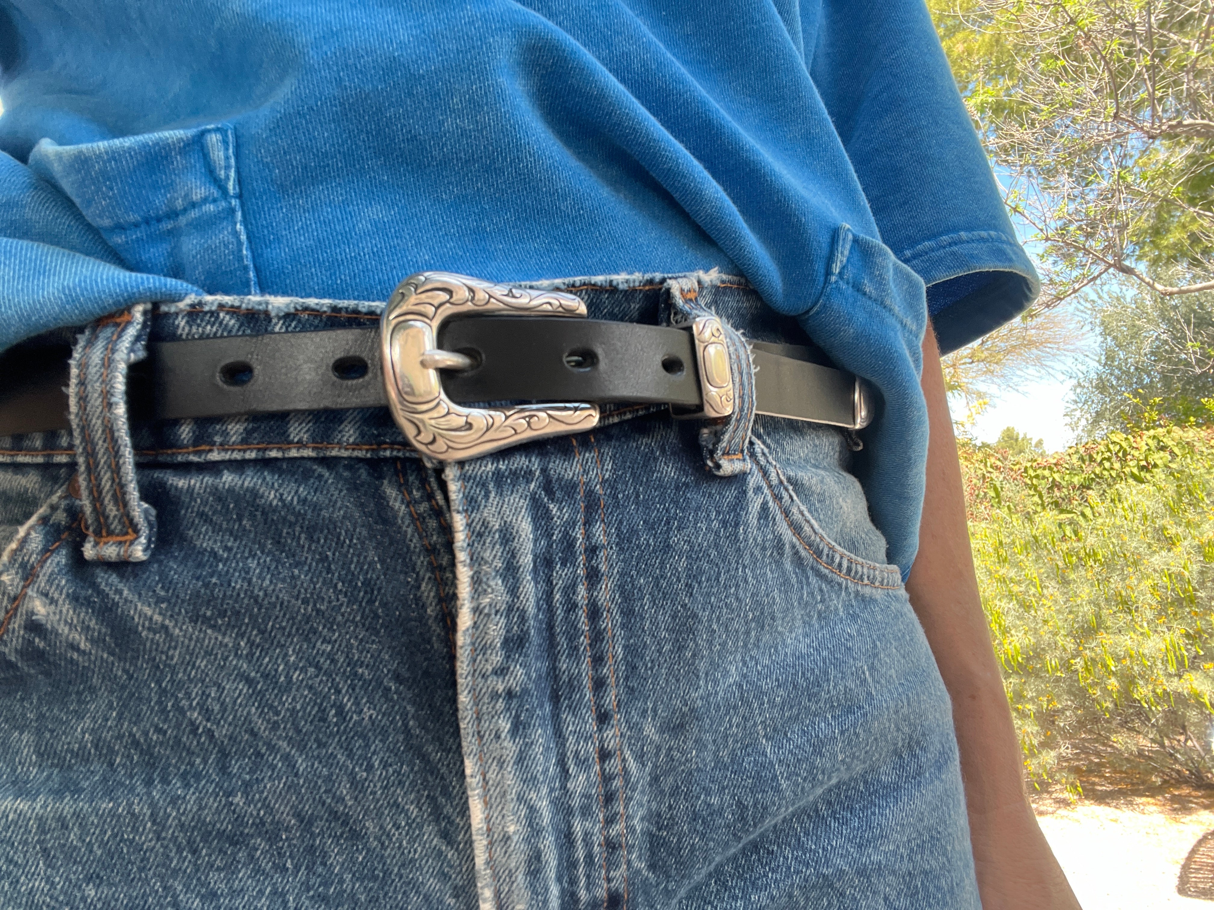 Person wearing blue jeans with a black belt and decorative buckle outdoors.