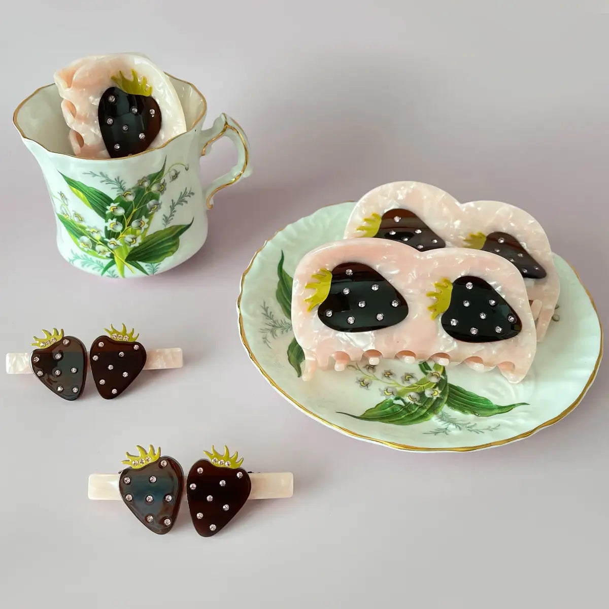 A few Centinelle, strawberries styled hair barrettes in a teacup and on a plate on a grey background