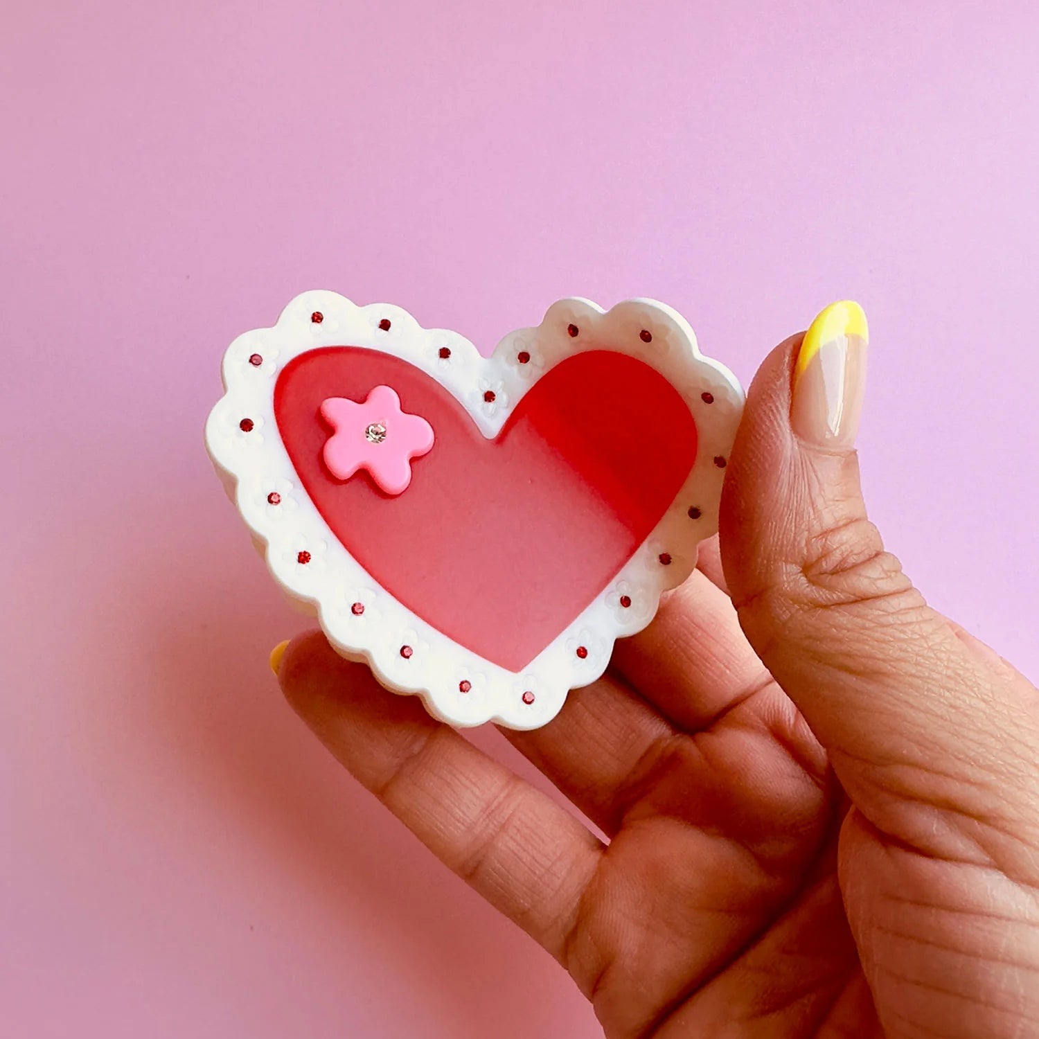 Close up of a person holding a Centinelle 'laced heart' style hair claw clip on a light pink background.