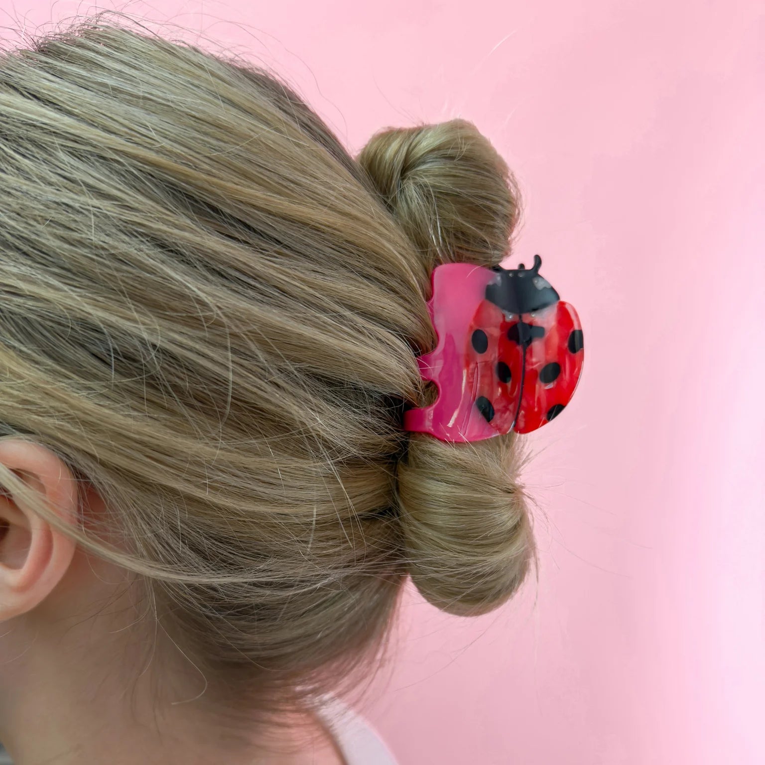 Pink, red, and black ladybug hair claw clip in a person's bonde hair. Light pink background