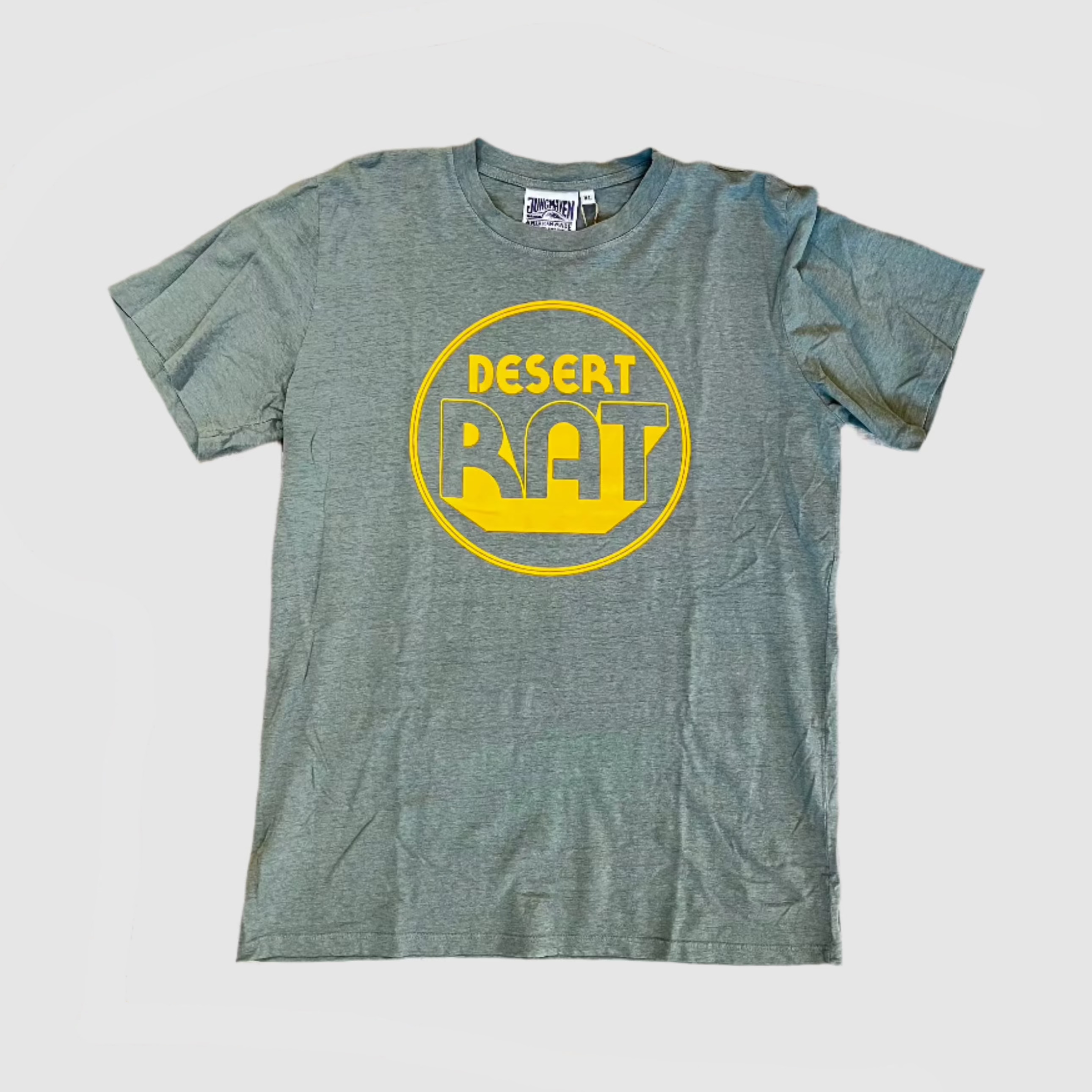 Front of a clay green colored t-shirt with "desert rat" circular, yellow colored logo centered on shirt. Gray background