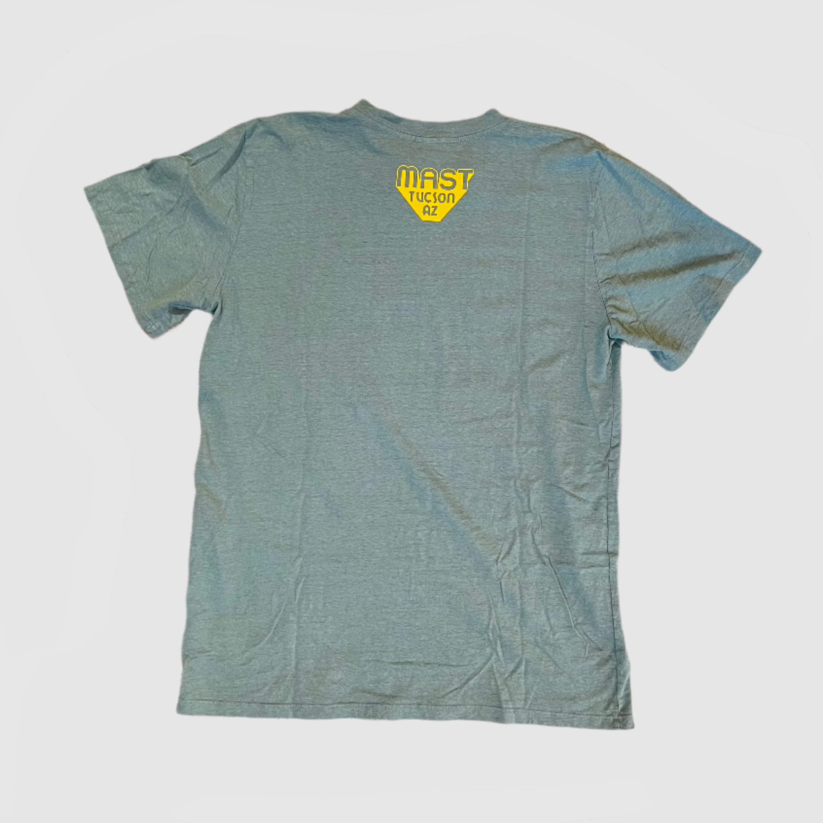 Back of clay green colored t-shirt with "MAST Tucson AZ" yellow colored logo centered towards top of shirt. Gray background