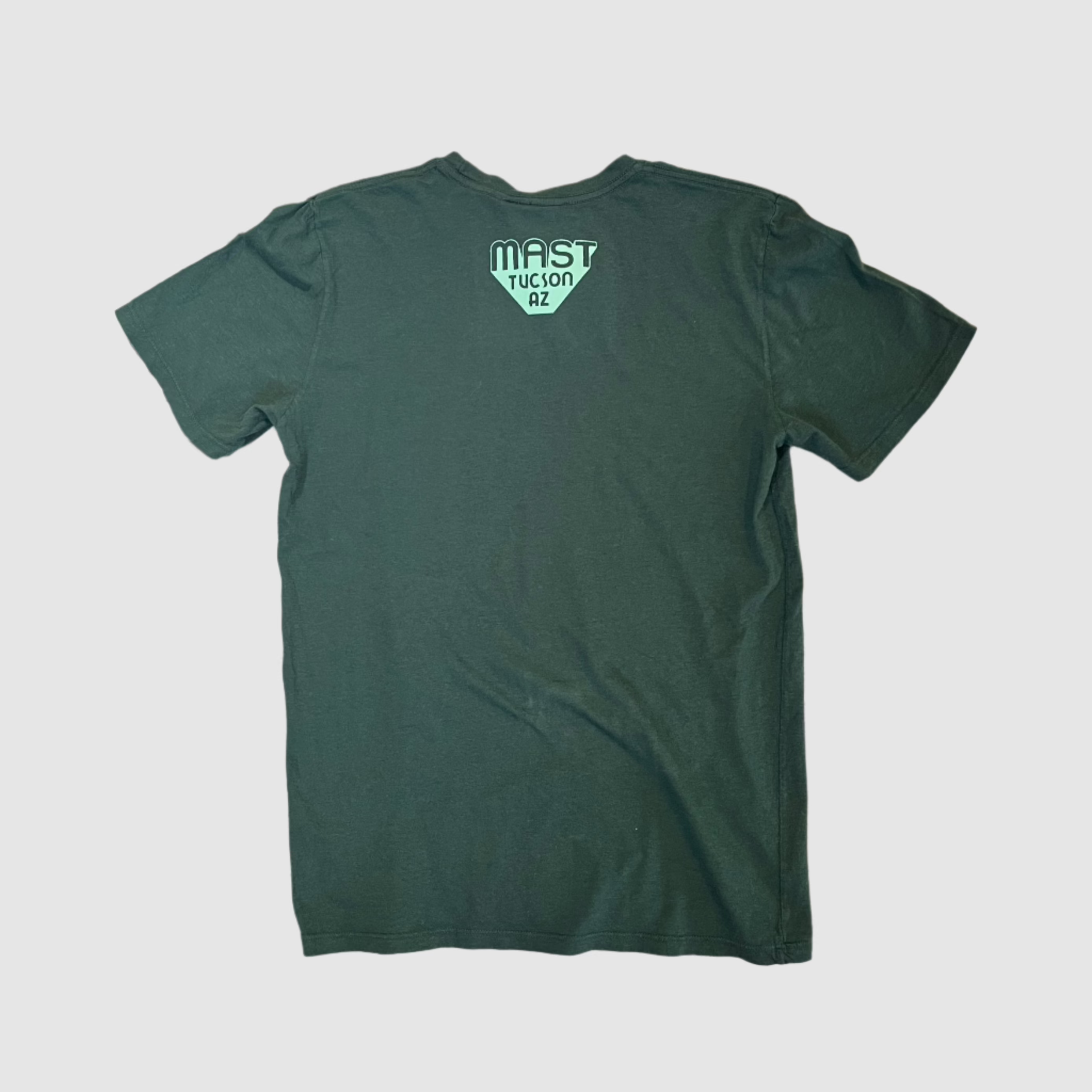 Back of pine colored t-shirt with "MAST Tucson AZ" mint colored logo centered towards top of shirt. Gray background