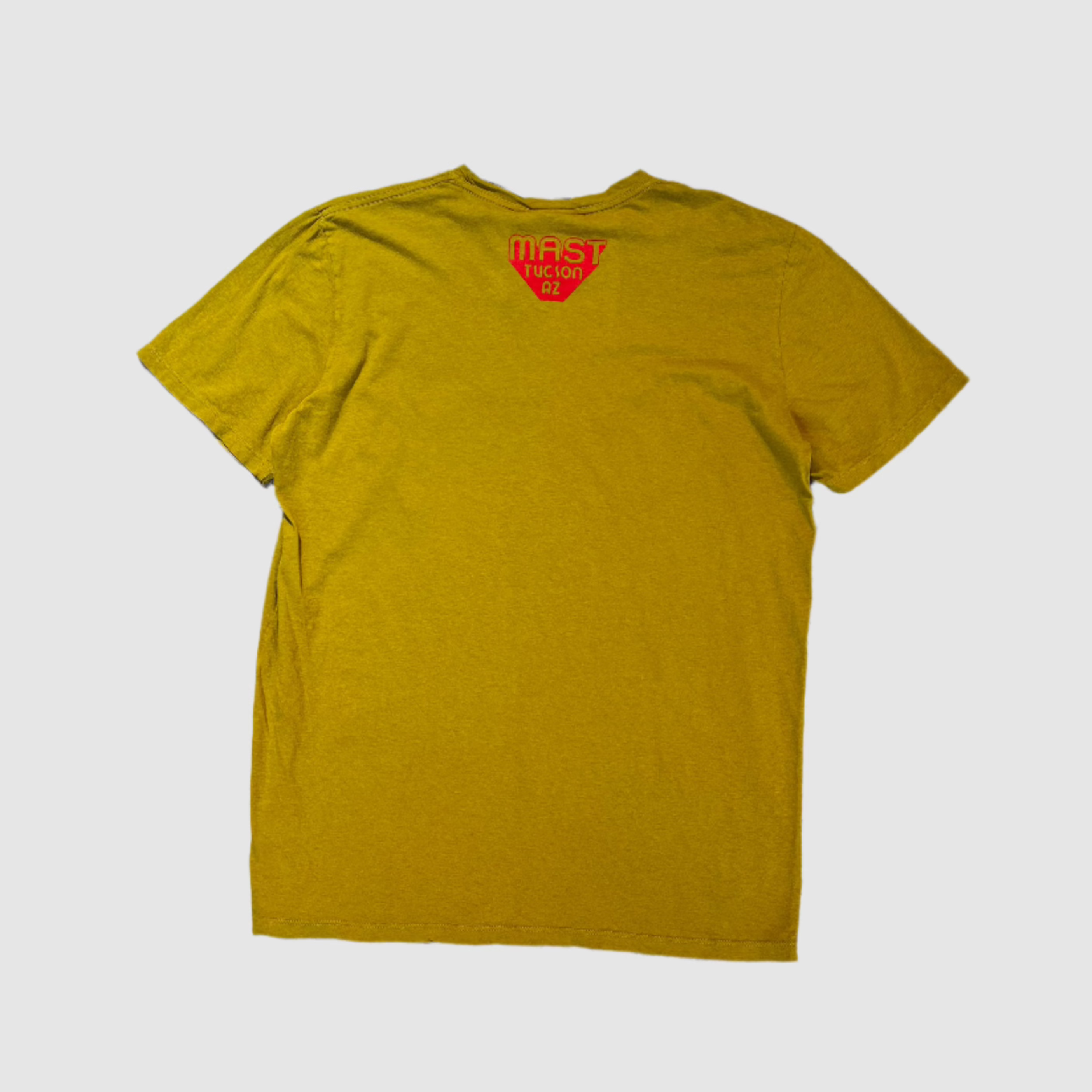 Back of a turmeric colored t-shirt with "MAST Tucson AZ" poppy colored logo in center towards top of shirt. On a gray background.