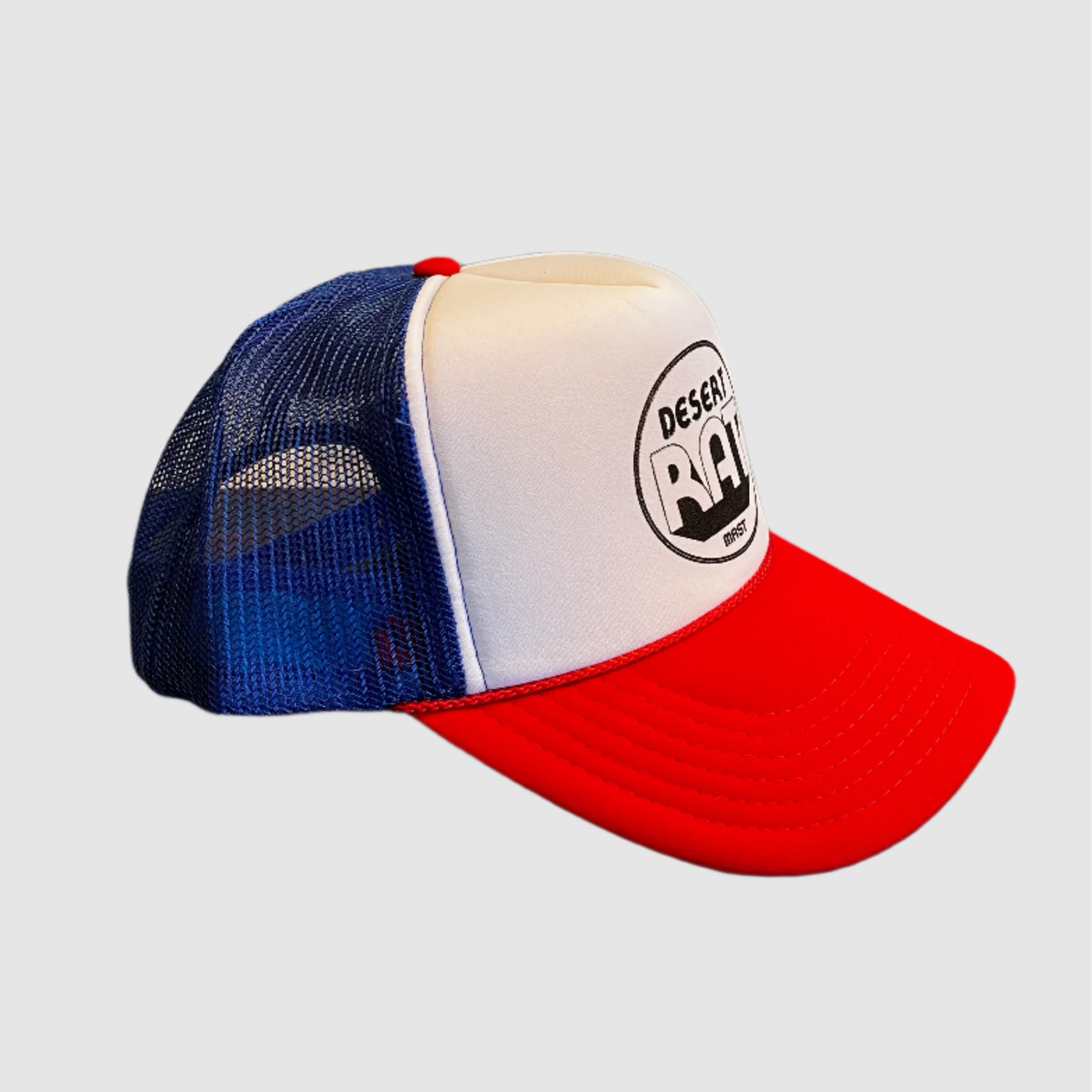 Side view of a "desert rat" logo printed in black on white trucker hat with red bill and blue mesh backing. Light gray background.