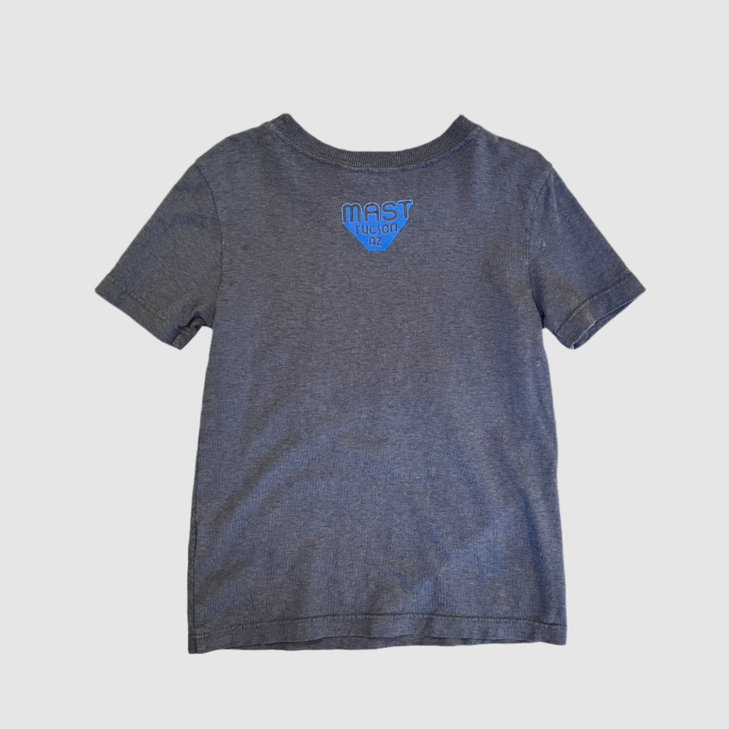 Back of a youth size, grey-blue colored t-shirt with "MAST Tucson AZ" neon violet colored logo centered towards top of shirt. Gray background