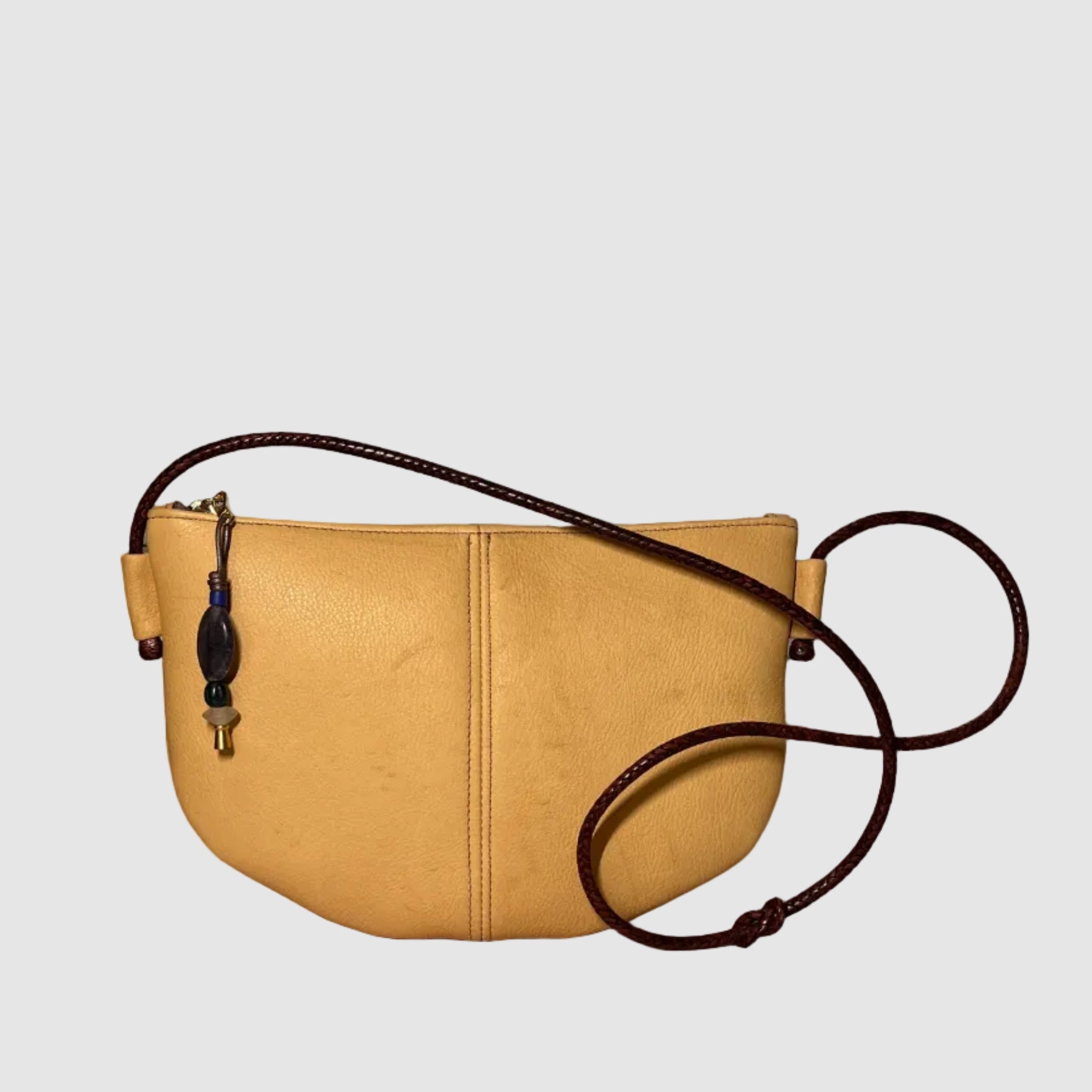 A matte, western, beige leather bag, handmade by Mellow Dawn in Tucson, Arizona. The bag features natural scarring and variation and a dark brown hand woven strap made in Mali. The bag is shown floating on a light grey background.