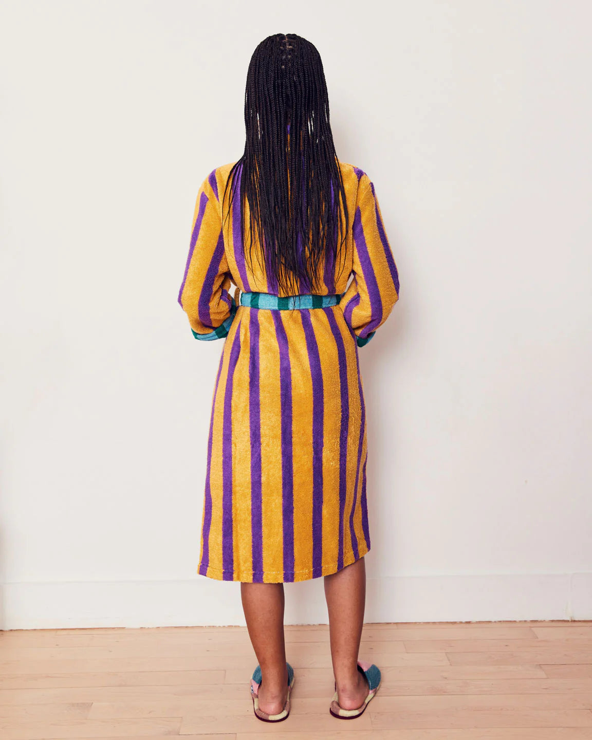 Person wearing a yellow and purple striped dress with a green belt against a white wall.