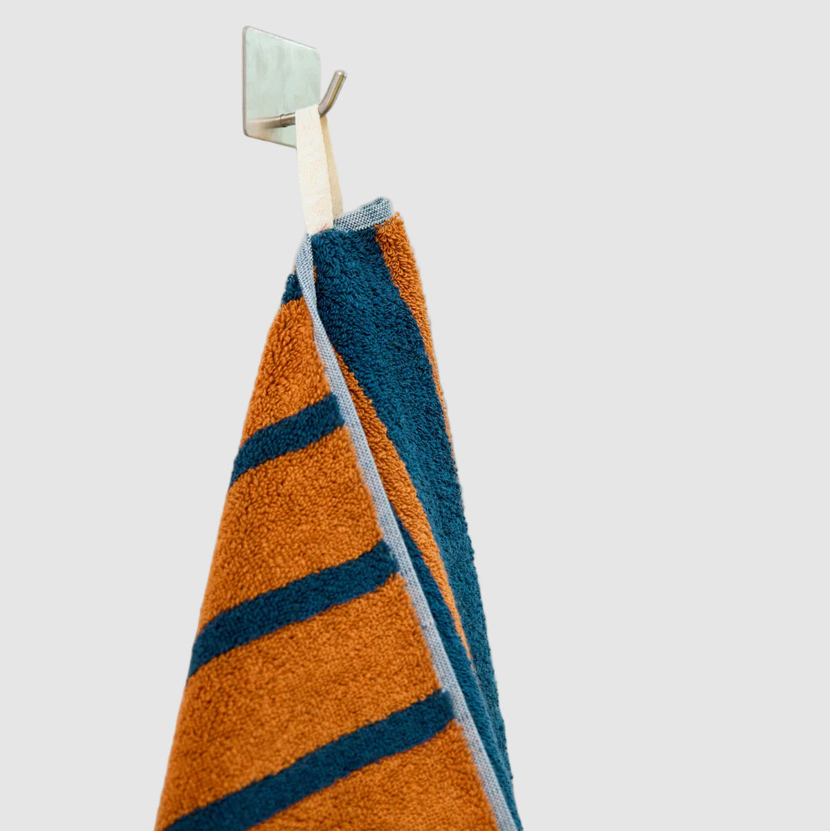 Striped dusen dusen hand towel with orange and blue striped pattern hanging on a wall hook on a light grey "wall" background