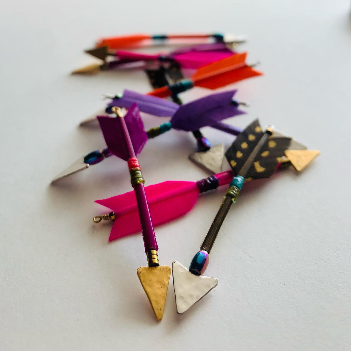 Group of multicolored feather arrow earrings with vermeil and silver arrowheads and leverback clasps designed and handmade in Tucson, Arizona by Sofie Albertsen. Displayed on a light grey background.