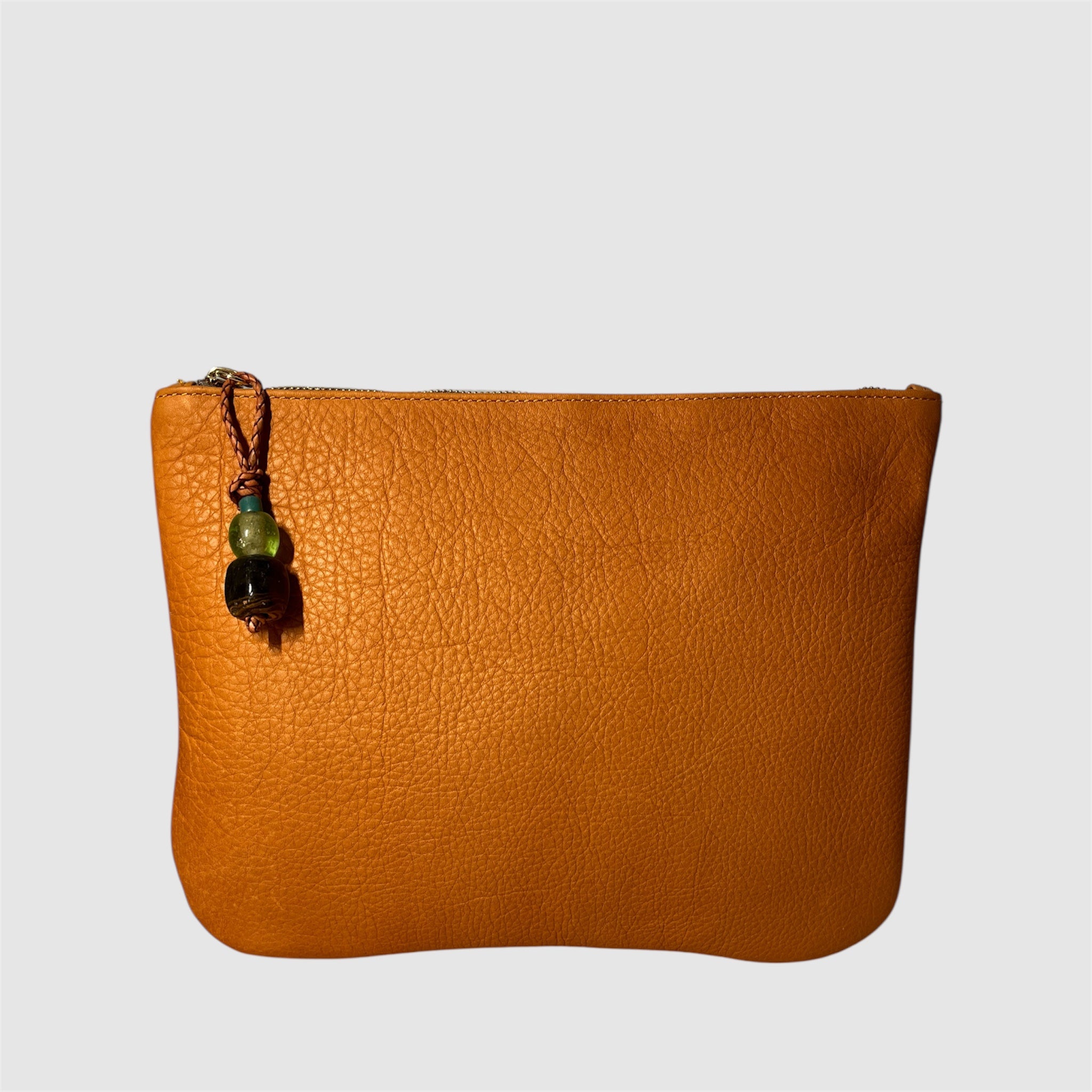 A tan leather clutch handmade by Mellow Dawn in Tucson, Arizona. The bag features natural scarring and variation, a glass bead zipper pull with braided leather attachment. The clutch is shown floating on a light grey background
