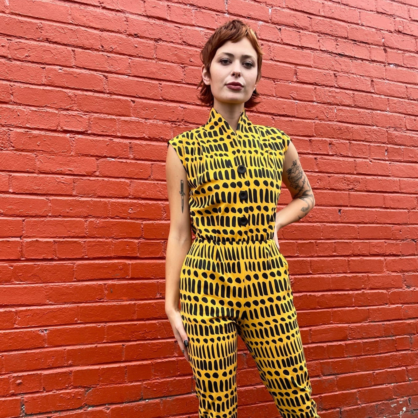 Noowork's "Dorcas Code" pattern featuring a yellow background and various black stripes and dots printed on a cotton "magic suit" styled on a young person. Reddish brick background.
