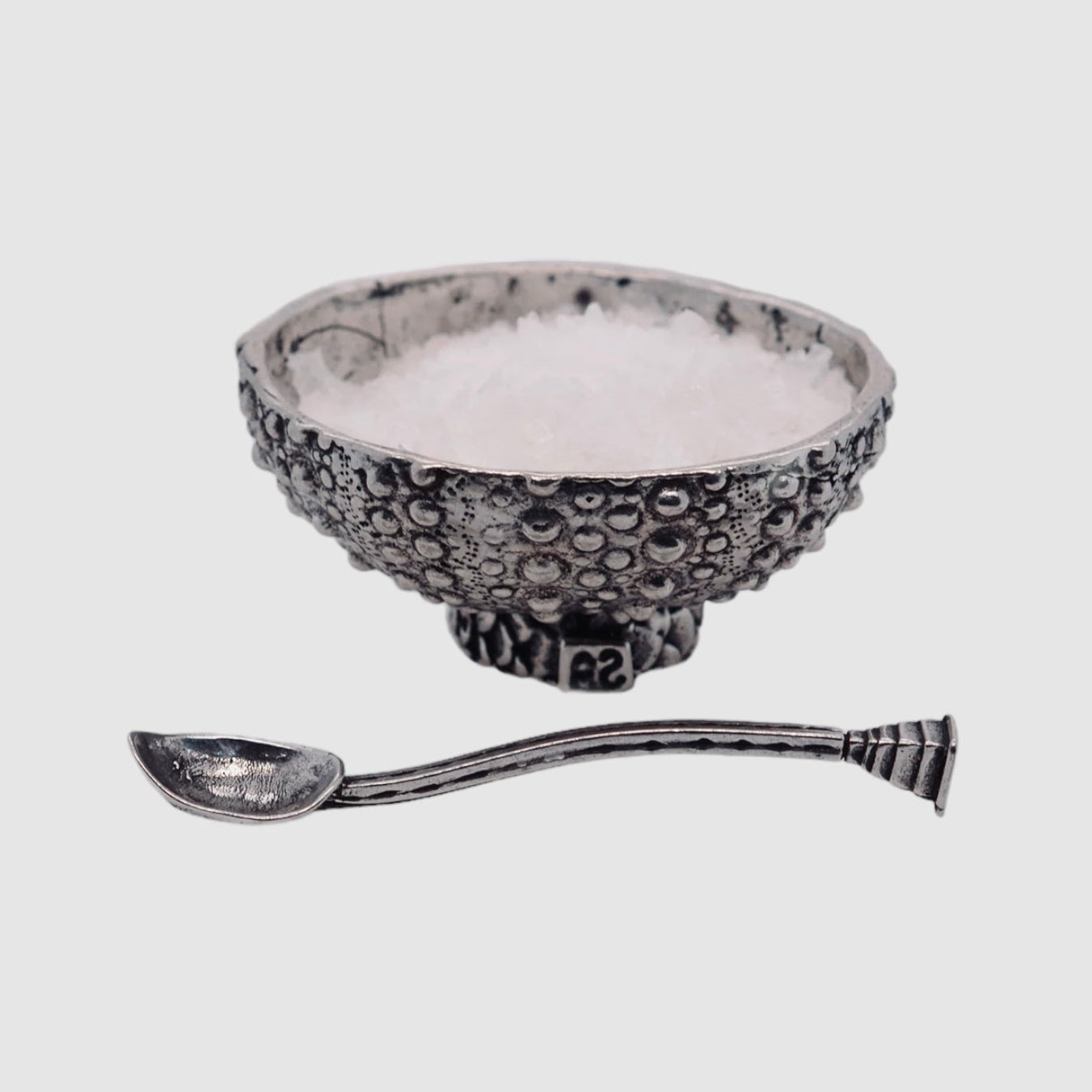 A pewter bowl with a sea urchin pattern accompanied by a mini spoon, both items are silver in color.