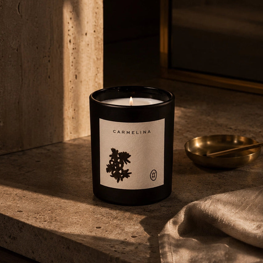 A black glass candle jar labeled 'CARMELINA' with a white botanical label, placed on a stone countertop in warm sunlight beside a folded linen cloth and a small brass dish.