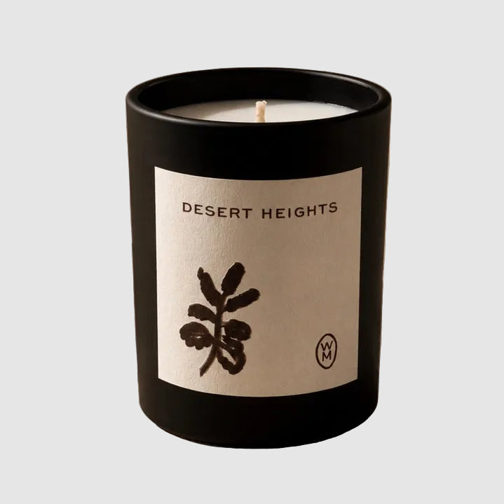 A black glass candle jar labeled 'DESERT HEIGHTS' sits on a textured stone surface beside a large rock, with warm sunlight casting shadows across the scene.