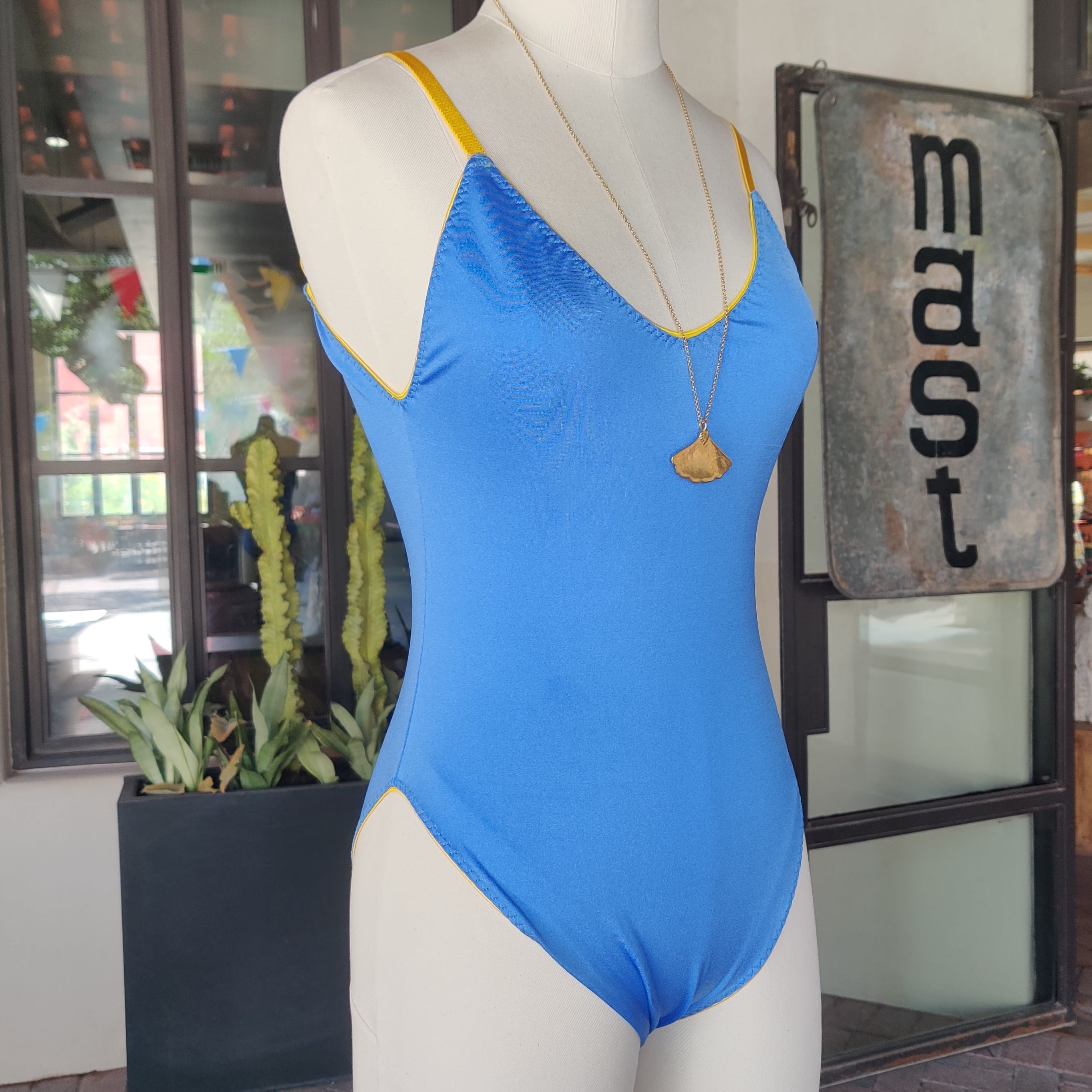 A bright blue one-piece swimsuit with yellow trim displayed on a white mannequin torso, set against a storefront background.