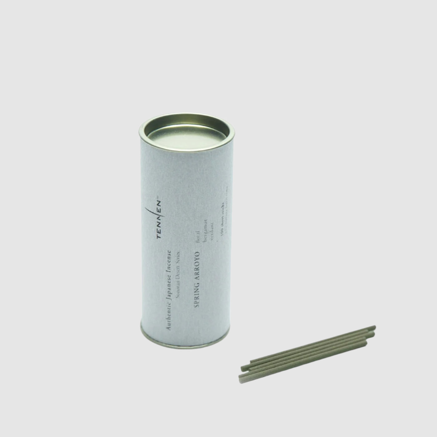 Canister of Tennen Spring Arroyo Incense next to a few loose sticks of incense on a grey background. 