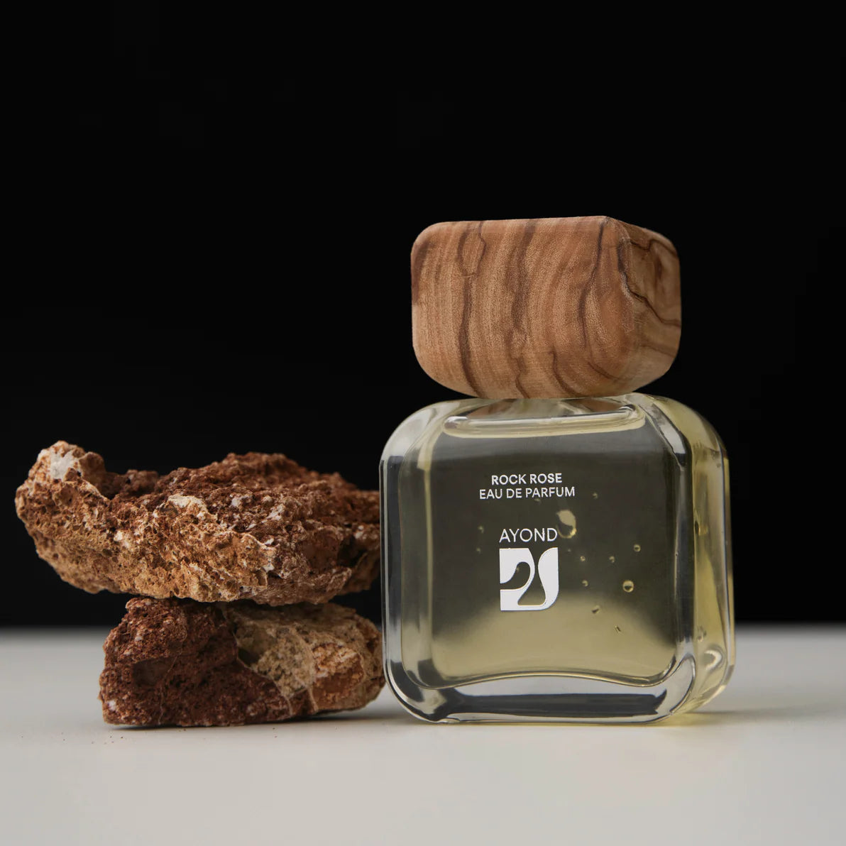 Perfume bottle with wooden cap and 'Ayond' branding next to rocks on a dark background