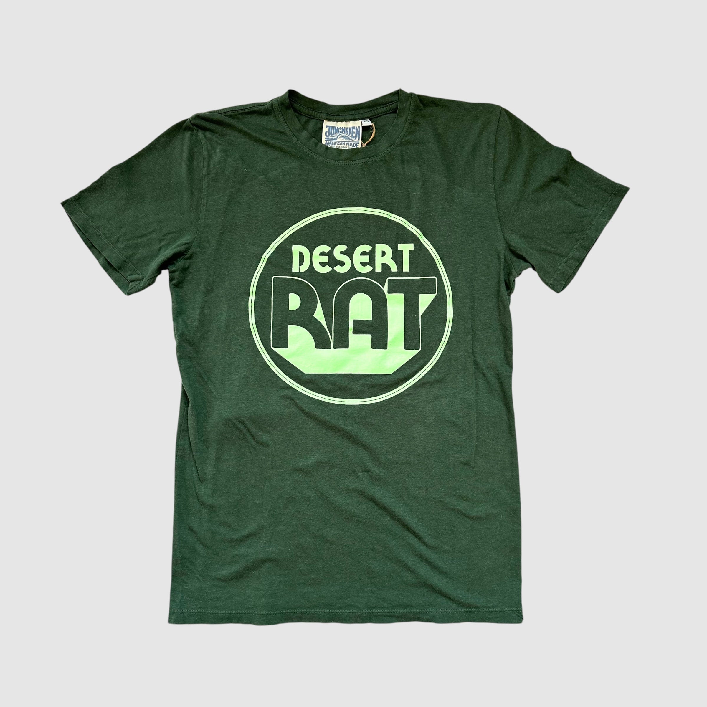 Front of a pine colored t-shirt with "desert rat" circular, mint colored logo centered on shirt. Gray background