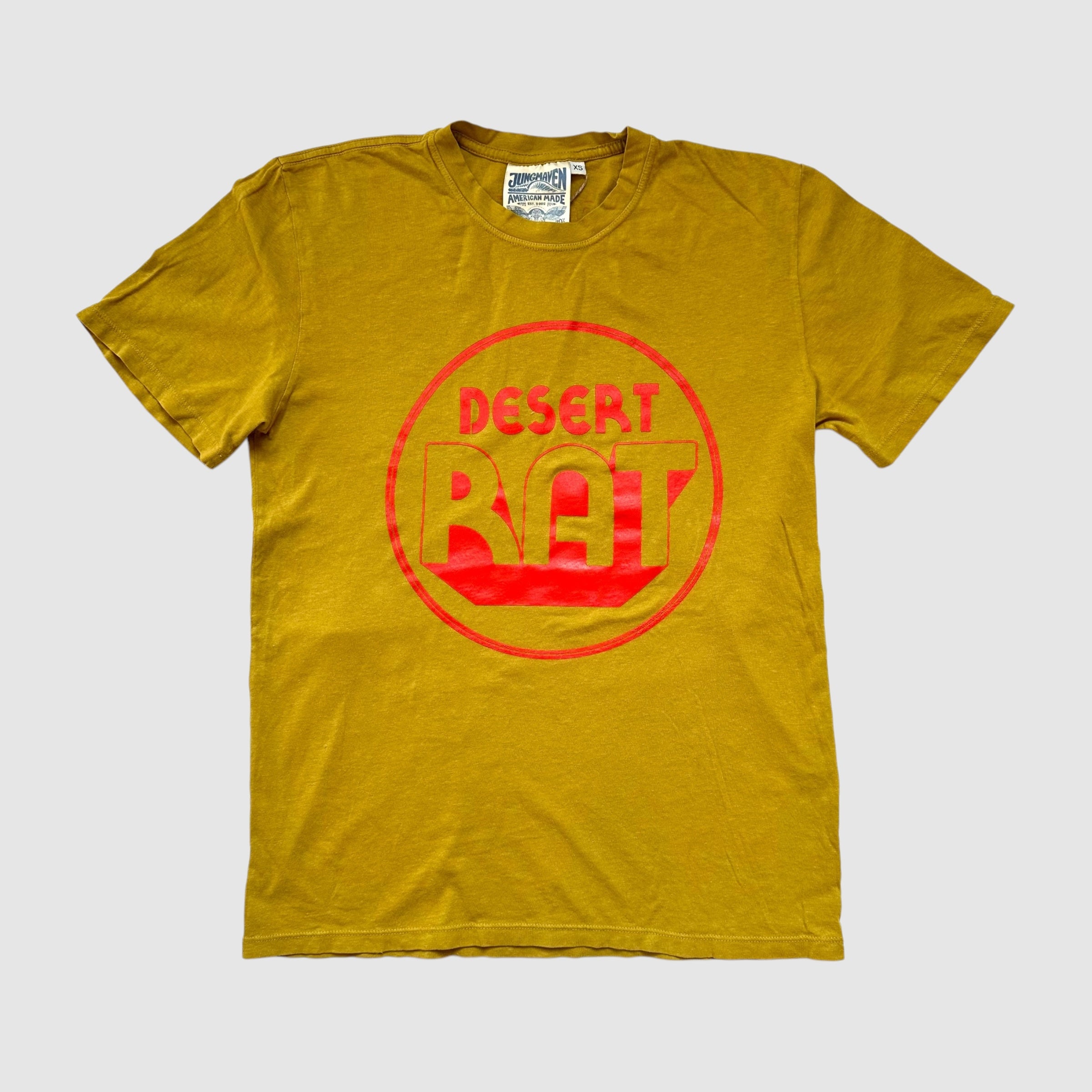 Front of turmeric colored t-shirt with "desert rat" circular, poppy colored logo centered. On a gray background.