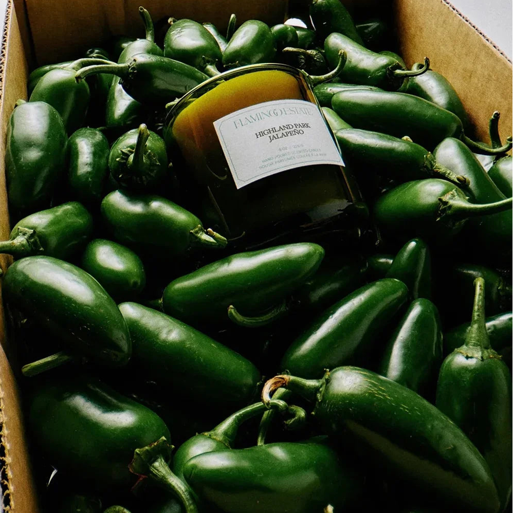 "Highland Park Jalapeño" scented Flamingo Estate Candle surrounded by jalapeño peppers in a cardboard box