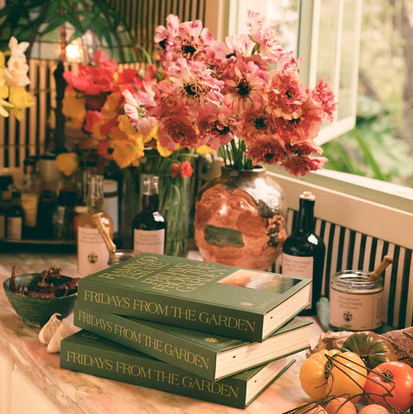 Stack of books titled 'Fridays from the Garden' on a table with flowers and fruits.