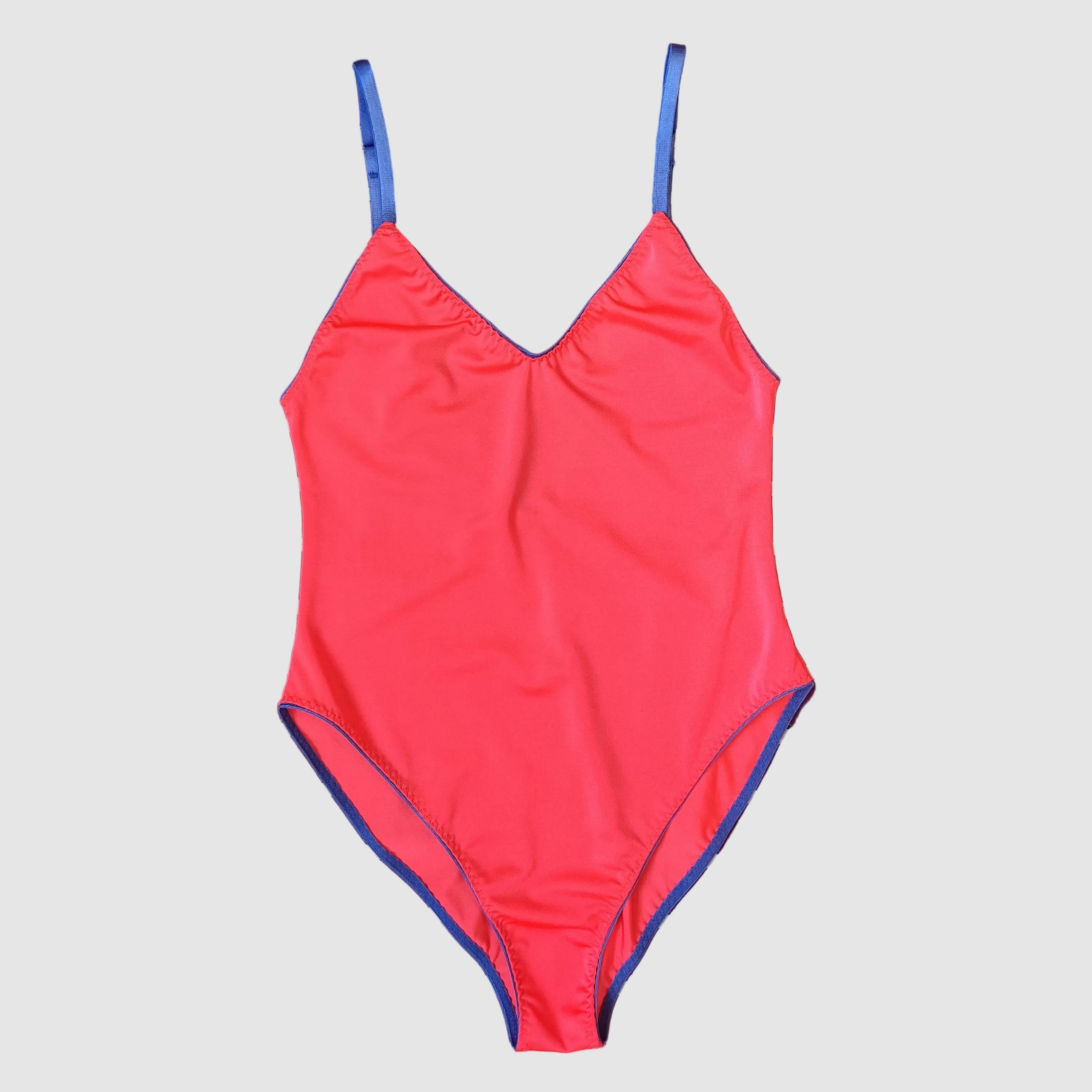 A bright coral pink one-piece swimsuit with blue trim and matching blue straps, laid flat on a dark purple and white striped towel.