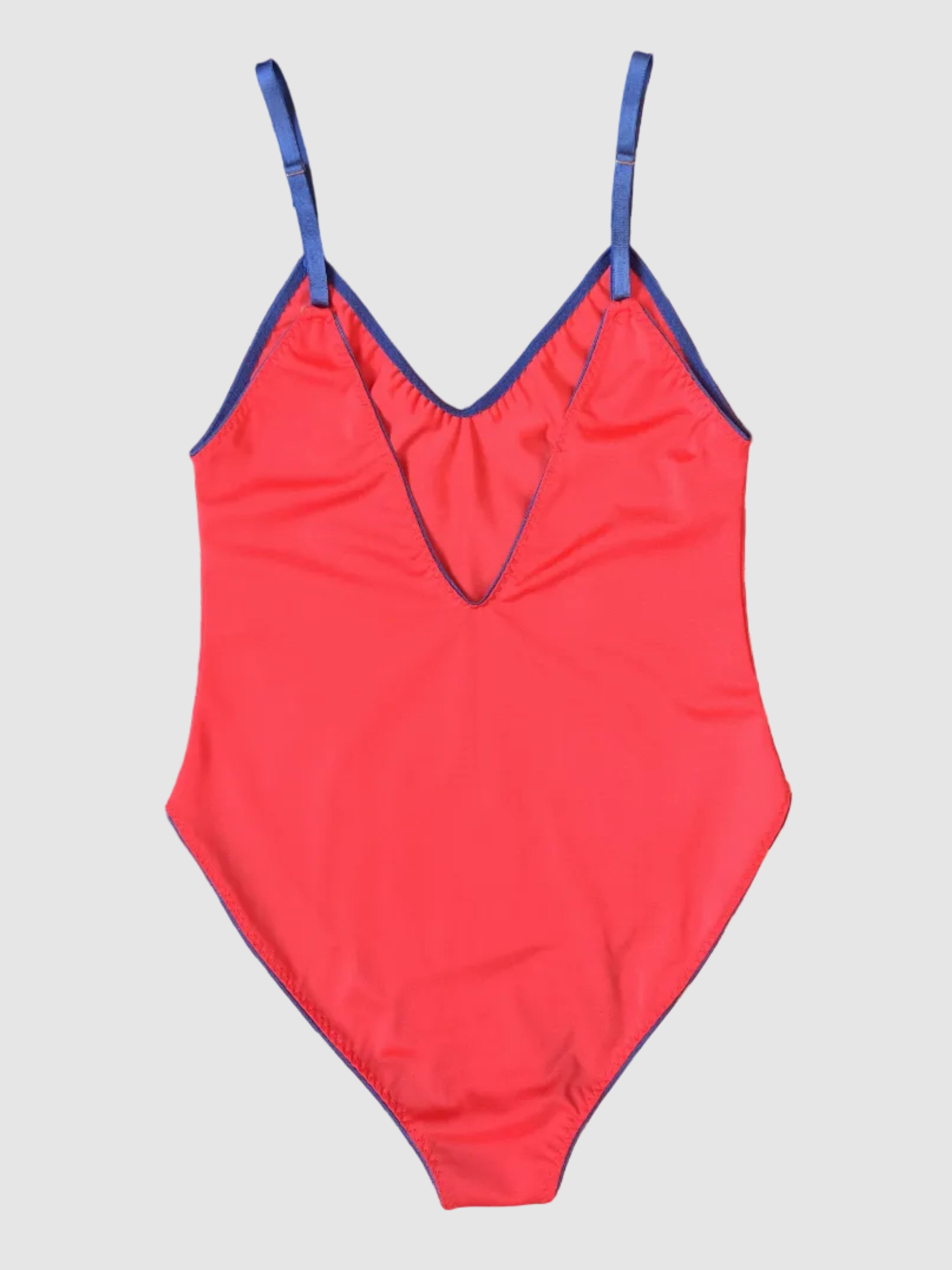 A bright coral-red one-piece swimsuit with blue trim and shoulder straps, laid flat on a dark purple and white striped fabric background.