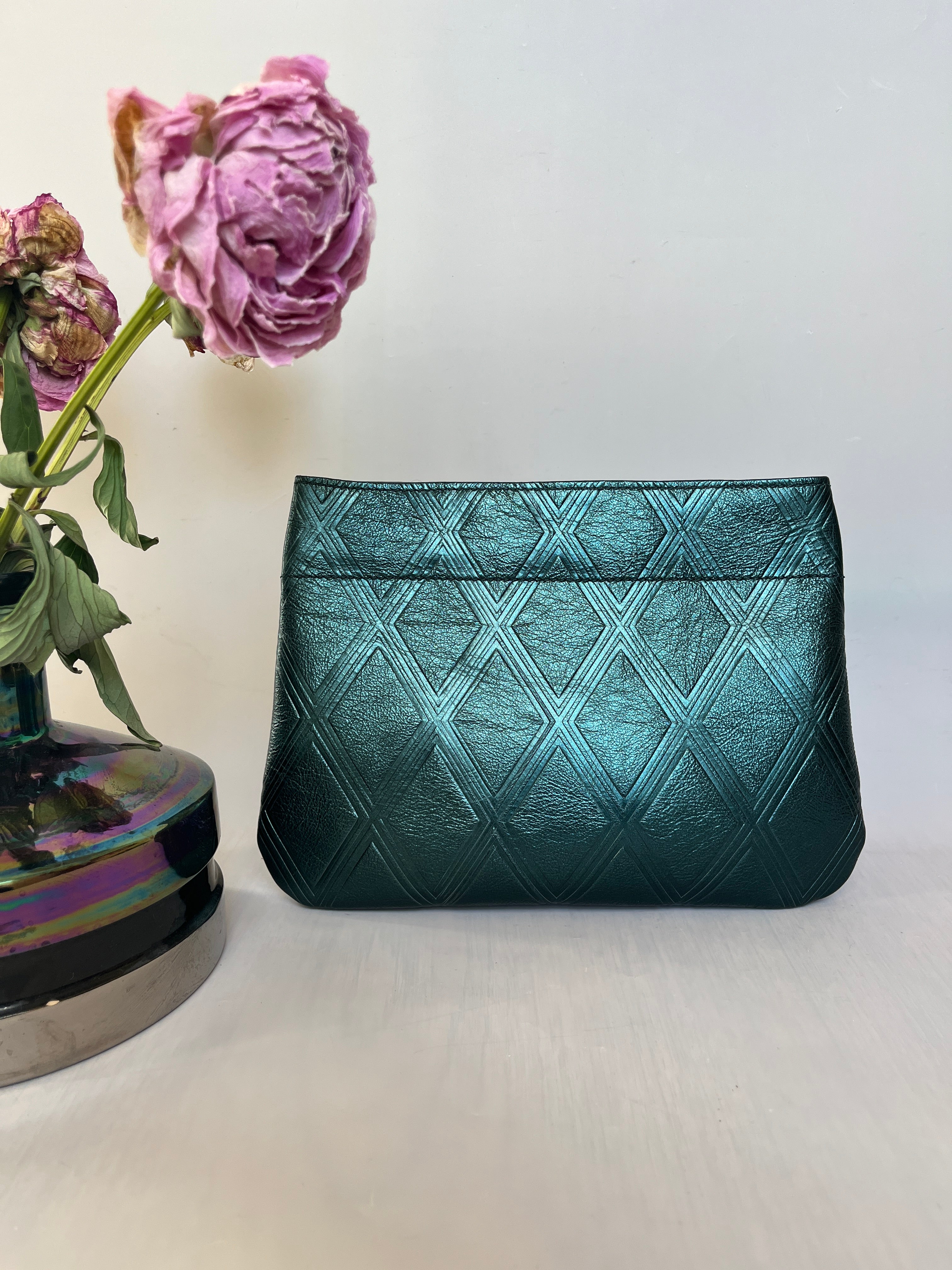 Embossed metallic green handmade leather clutch next to a vase of flowers on a light grey background. Bag made by Mellow Dawn in Tucson, Arizona.