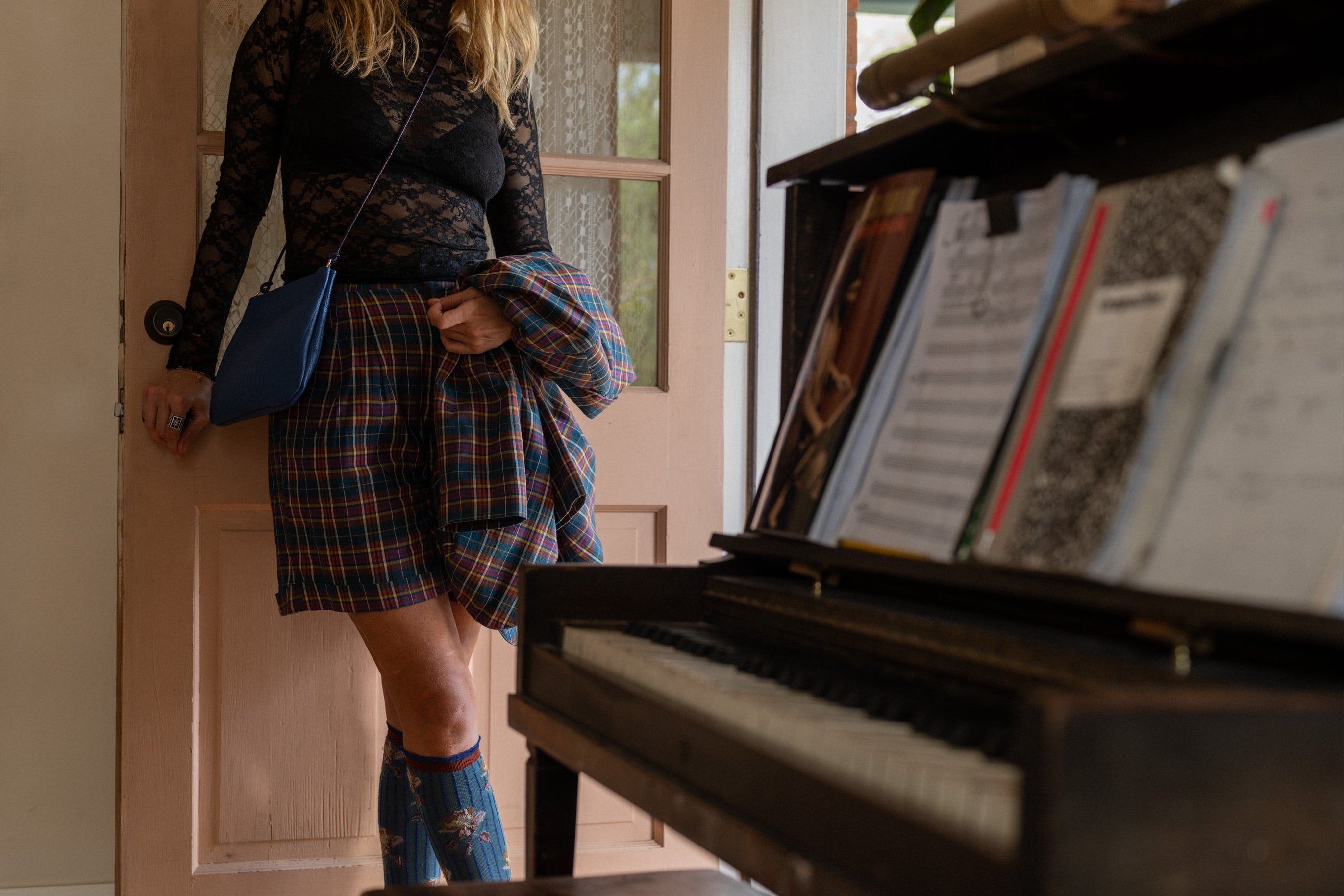 Woman standing next to a piano wearing a plaid outfit and an embossed cobalt blue handmade leather handbag with twisted leather strap made by Mellow Dawn in Tucson, Arizona. 
