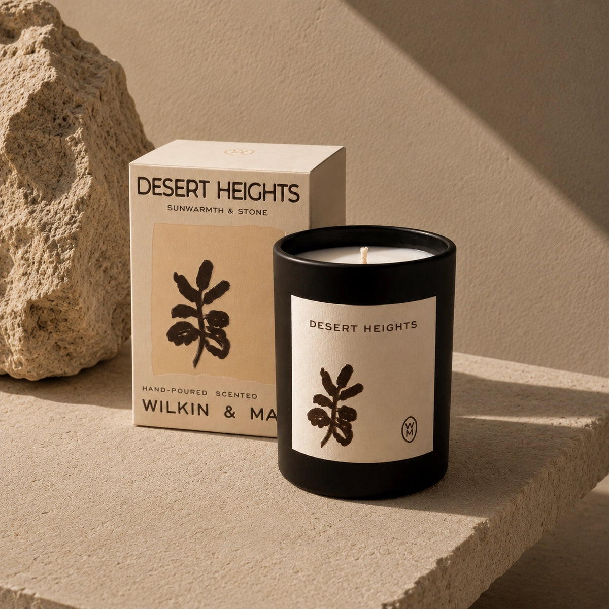 A black glass candle jar labeled Desert Heights sits on a stone surface beside its cream-colored product box. The label shows a dark botanical leaf illustration, and the box reads Desert Heights, Sunwharm & Stone, Hand-Poured Scented, Wilkin & Mar.