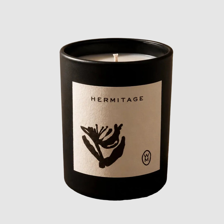 A black glass jar candle with a white label reading HERMITAGE and a black botanical illustration, placed on a rustic wooden surface against a textured beige wall with soft leaf shadows.