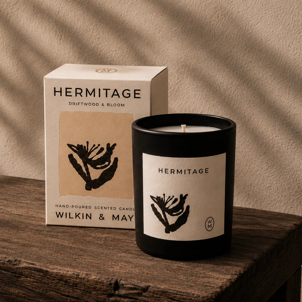 A Hermitage scented candle in a matte black jar with a cream label sits on a rustic wooden table next to its cream-colored product box. The background is a textured beige wall with soft leaf shadows cast across it.