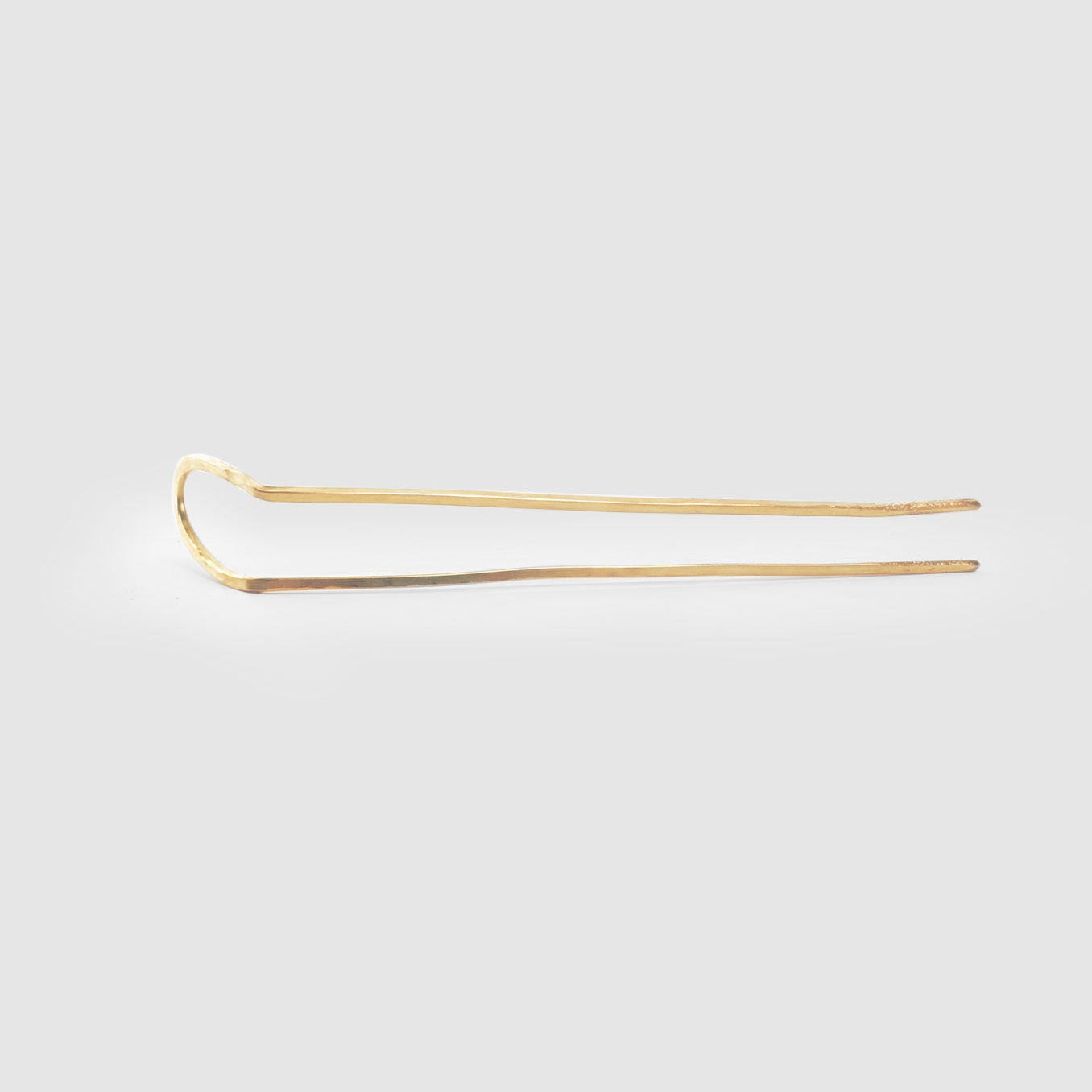 A long length hand hammered brass hairpin from Tasha Bundy's ARCO collection on a light grey background.